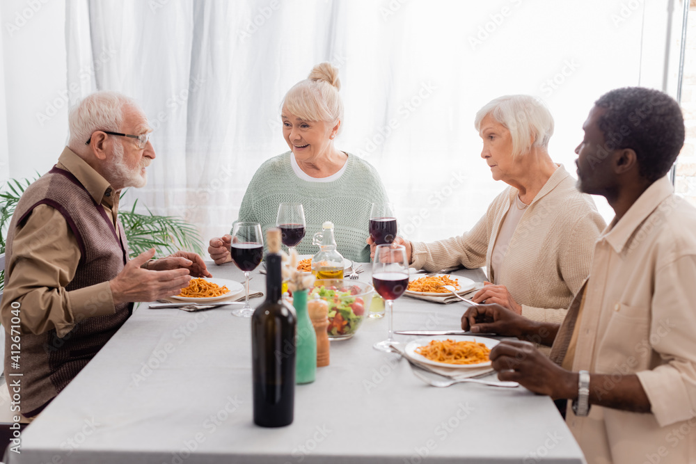 happy multicultural and senior friends talking while having delicious lunch near glasses of red wine happy multicultural and senior friends talking while having delicious lunch near glasses of red wine