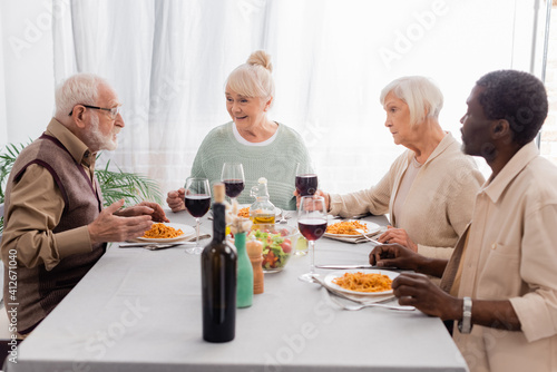 happy multicultural and senior friends talking while having delicious lunch near glasses of red wine