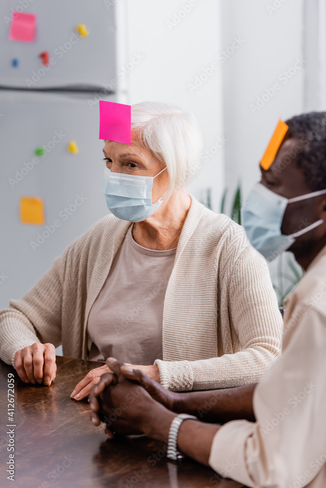 multicultural senior friends in medical masks with sticky notes on foreheads playing game in kitchen multicultural senior friends in medical masks with sticky notes on foreheads playing game in kitchen