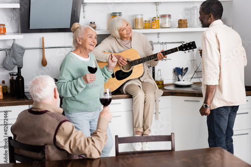 smiling senior woman playing acoustic guitar near retired multicultural friends in kitchen