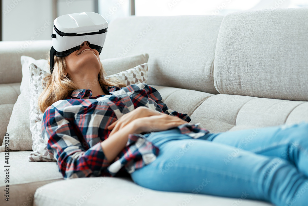 Obraz premium Beautiful young woman using virtual reality headset and enjoying the moment lying on couch at home.