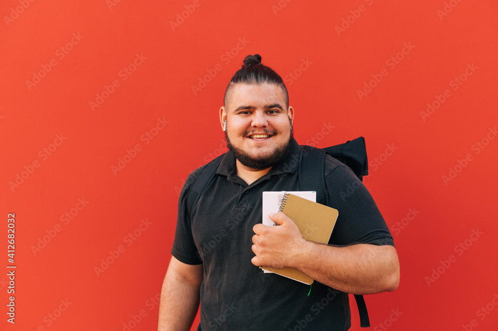 Portrait of happy overweight student on red background, wearing dark clothes, standing with books and notebook in hands and backpack on his back on red background and smiling. Portrait of happy overweight student on red background, wearing dark clothes, standing with books and notebook in hands and backpack on his back on red background and smiling.