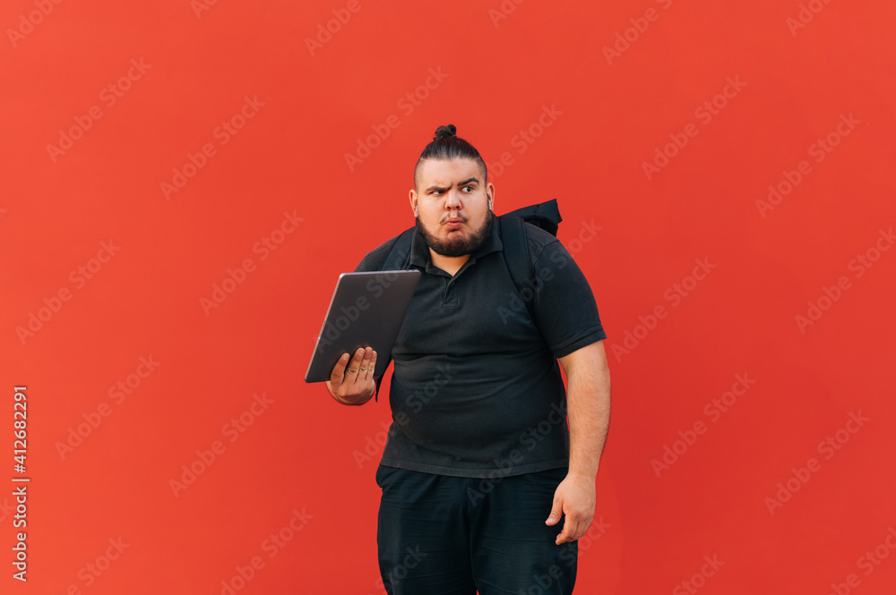 Funny overweight man in black clothes stands with a laptop on a red background and looks away in confusion, making a funny face. Funny overweight man in black clothes stands with a laptop on a red background and looks away in confusion, making a funny face.