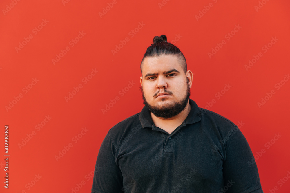 Portrait of a fat young man with a beard on a background of a red wall in a black T-shirt looking at the camera with a serious face. Portrait of a fat young man with a beard on a background of a red wall in a black T-shirt looking at the camera with a serious face.
