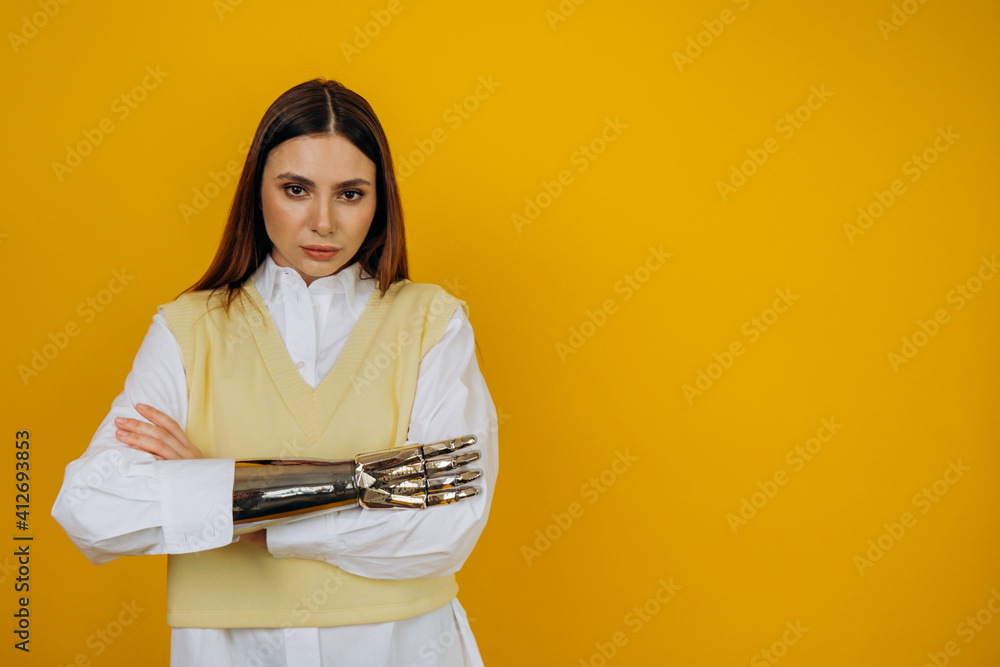 Portrait of a serious girl with a bionic prosthetic arm in a white shirt on a yellow background, hands folded at the waist in front of her and purposefully looking at the camera, copyspace. Portrait of a serious girl with a bionic prosthetic arm in a white shirt on a yellow background, hands folded at the waist in front of her and purposefully looking at the camera, copyspace.