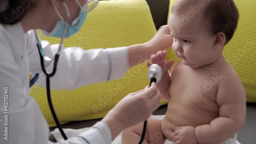 medicine and health, pediatrics, covid-19 concept - close-up young woman nurse or doctor pediatrician of Caucasian Slavic ethnicity examines infant 8-12 months baby on yellow gray sofa opposite window