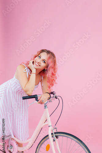 Refined woman in striped dress posing emotionally on a pink background. Gorgeous girl standing in studio with the bicycle.