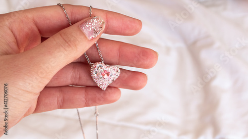 A beautiful pale pink heart pendant with a silver chain in a woman's hand. Fingernails with a silver glitter manicure, contrasted against a white bed spread in London, Ontario, Canada.