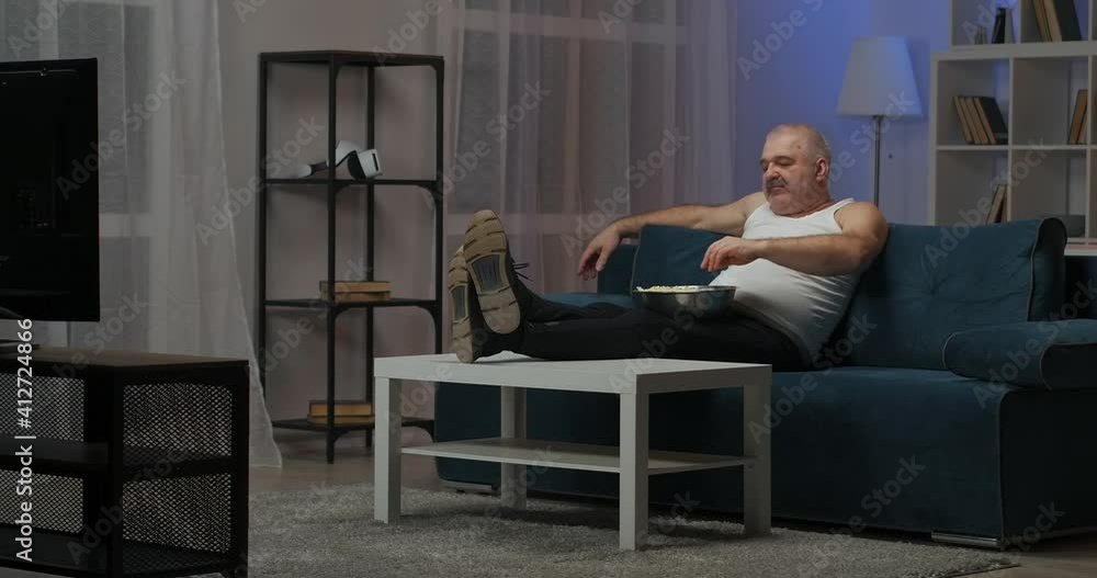 Tired man watching TV with a big bowl of popcorn. The old man watches TV at night, relaxing after a hard day. The man is sitting on the couch. Tired man watching TV with a big bowl of popcorn. The old man watches TV at night, relaxing after a hard day. The man is sitting on the couch.