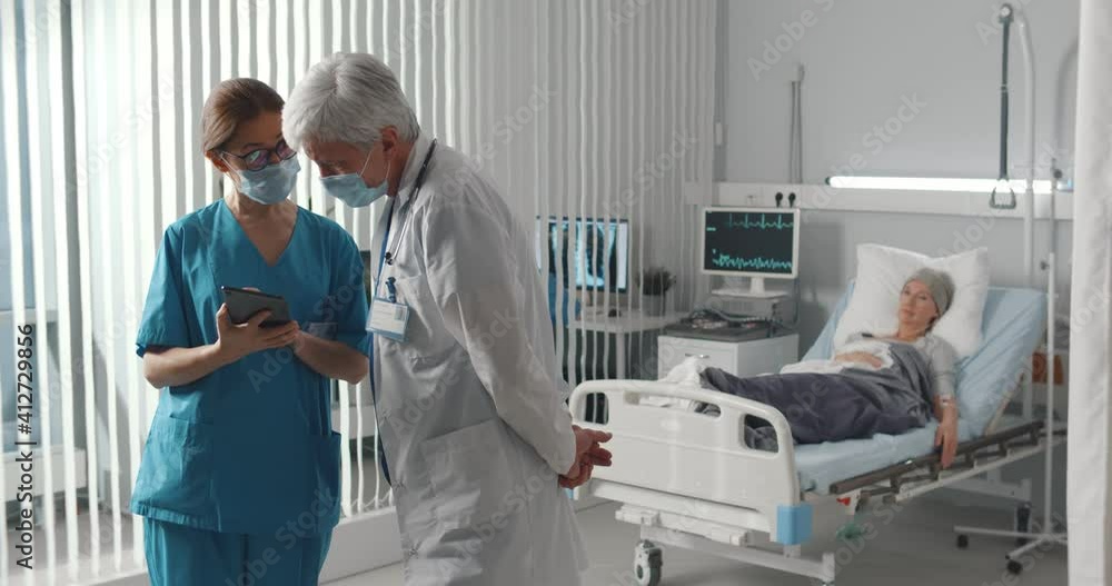Doctor and nurse in safety mask discussing diagnosis on tablet with woman patient lying in bed Doctor and nurse in safety mask discussing diagnosis on tablet with woman patient lying in bed