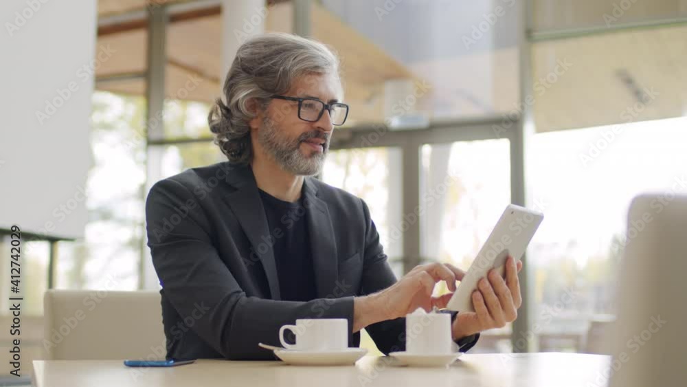 Medium shot of senior caucasian business man sitting in posh restaurant with cup of coffee smiling while using digital tablet Medium shot of senior caucasian business man sitting in posh restaurant with cup of coffee smiling while using digital tablet