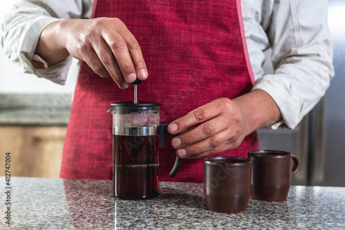 Close-up of a man's hands in a red apron pressing coffee in a French press