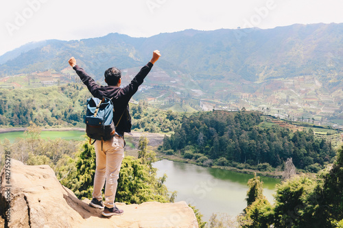 Young man standing on a rock of a cliff and enjoying the view of nature of lake and mountain in Dieng Indonesia.