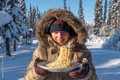 Woman standing outdoors in northern cold arctic landscape during freezing -40 temperature day holding a plate of frozen pasta noodles with fork holding up stiff food. Wearing beige, faux fur jacket.  photo