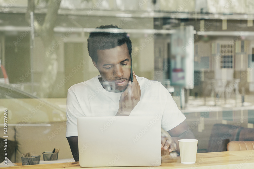 Serious African American man typing on laptop and speaking on cellphone in co-working space. View from street through window glass. Communication concept Serious African American man typing on laptop and speaking on cellphone in co-working space. View from street through window glass. Communication concept