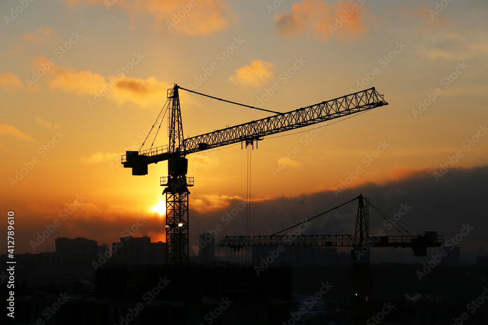 Silhouettes of construction cranes and unfinished residential buildings on sunrise background. Housing construction, apartment block in winter city