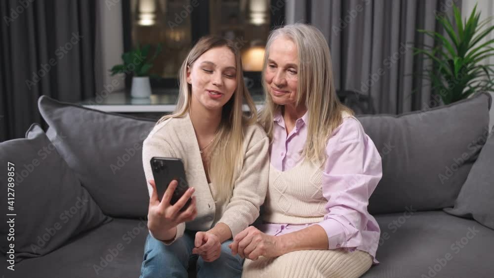Granddaughter and grandmother view photos on a smartphone sitting in the living room, family day, positive emotions. Granddaughter and grandmother view photos on a smartphone sitting in the living room, family day, positive emotions.