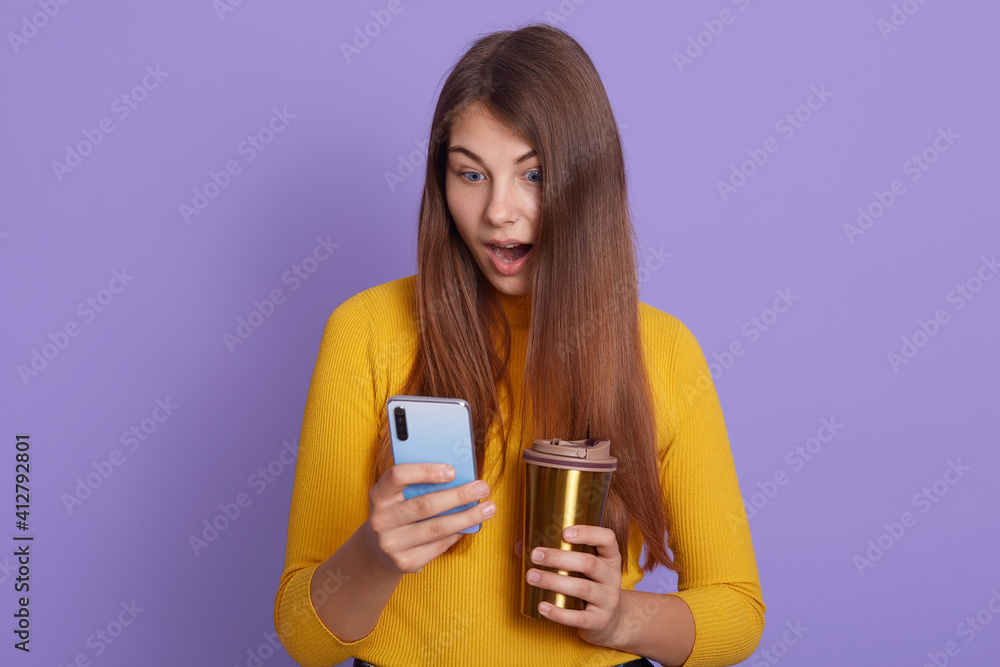 Photo of surprised woman being surprised, shocked to see new post, connected to wireless internet, reads astonishing news, drinks coffee or tea from thermo mug, wearing yellow jumper.