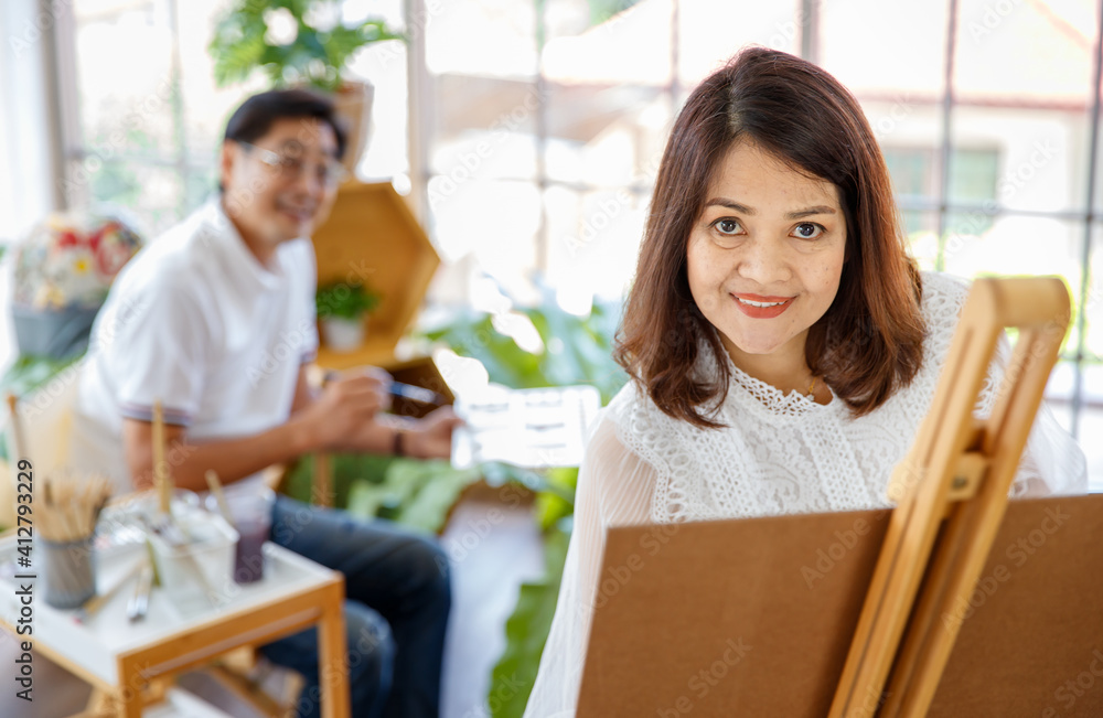 Senior woman painting image in studio with husband blur in background. Senior woman painting image in studio with husband blur in background.
