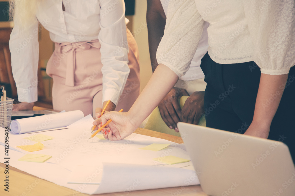 Unrecognizable designers drawing on big sheet of paper and sharing ideas. Woman holding pencil and writing notes for project. Colleagues standing near table. Communication, design and teamwork concept