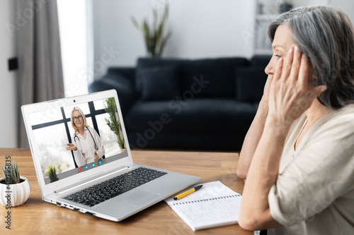 Senior mature woman sits at the table at home have video call with doctor, using virtual connection on laptop, female patient talks to webcam, consults with female physician on computer remotely © Vadim Pastuh