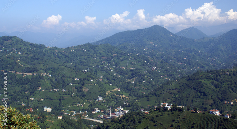 A panoramic shot of a scenery that captures valleys filled with green trees and houses under the cloudy, blue sky.