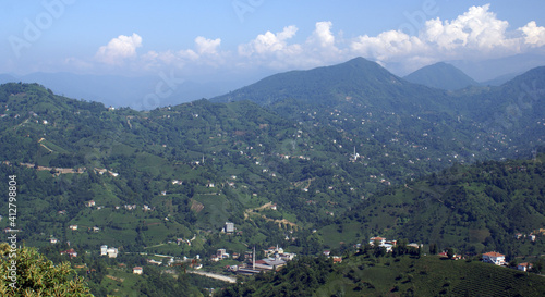 A panoramic shot of a scenery that captures valleys filled with green trees and houses under the cloudy  blue sky.