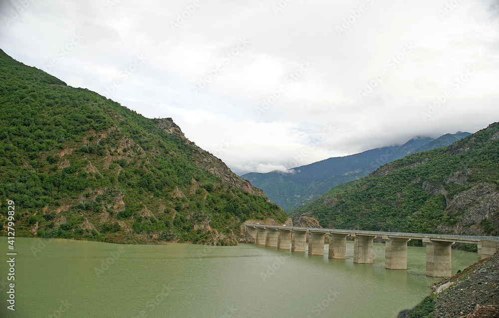 A panoramic shot of a scenery that captures valleys full of green grass, trees near a lake under the cloudy, blue sky.