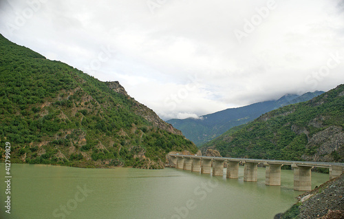 A panoramic shot of a scenery that captures valleys full of green grass, trees near a lake under the cloudy, blue sky.