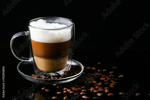 Cappuccino coffee in a transparent cup on a black background and scattered coffee beans.