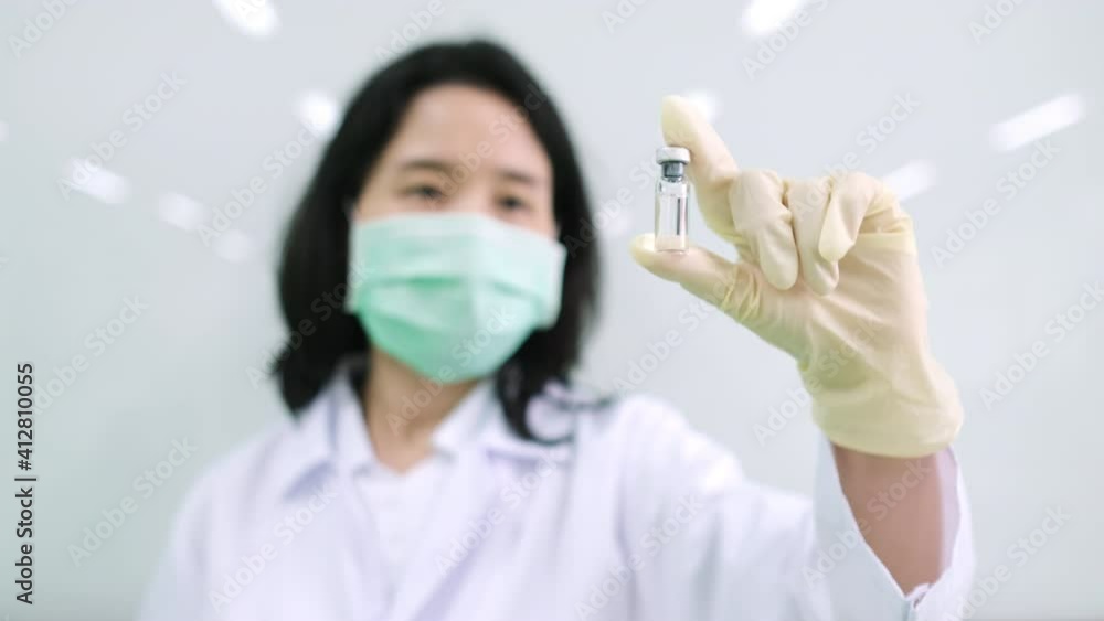 Scientist wearing medical mask and lab coat holding COVID vaccine vial on hand. Doctor looking at the bottle, success on the develop coronavirus vaccine. Focus on the vaccine vial. Research concept Scientist wearing medical mask and lab coat holding COVID vaccine vial on hand. Doctor looking at the bottle, success on the develop coronavirus vaccine. Focus on the vaccine vial. Research concept