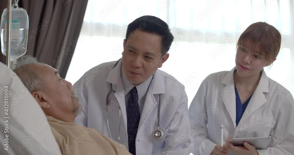 Asian male and female doctors come to talk, ask questions and take notes in tablet with recovering older man patient who lying in bed at hospital ward. Senior people healthcare support concept. Asian male and female doctors come to talk, ask questions and take notes in tablet with recovering older man patient who lying in bed at hospital ward. Senior people healthcare support concept.