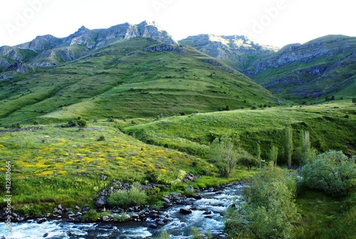 There is a valley filled with green grass and yellow flowers near a river.  © photograzon