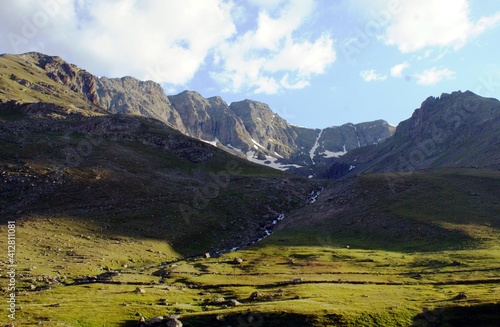 A panoramic  detailed shot of a mountain covered up with snows near a path filled with green grass and stones under the blue sky.