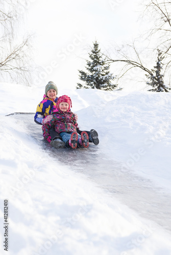 Winter activities outdoors. Cute happy little girls wearing a warm clothes are having fun, sliding down a hill on the ice slide, vertical