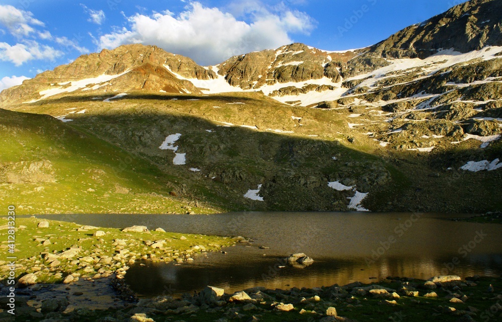 A panoramic shot of valleys which have snows on them while being covered with green grass and stones under the blue sky with a stream.