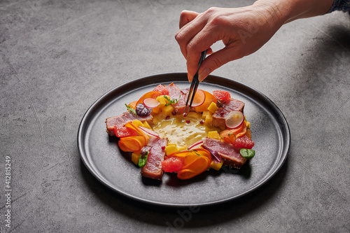 Woman's hand lays out the grain of red pepper with tweezers to homemade salad with tuna steak, mango sauce, herbs and spices on a dark plate, close up, food decorating concept