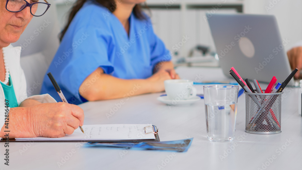 Close up of elderly woman doctor taking notes on clipboard while coworkers discussing in background, writing on laptop during brainstorming. Profesional teamworker having medical meeting Close up of elderly woman doctor taking notes on clipboard while coworkers discussing in background, writing on laptop during brainstorming. Profesional teamworker having medical meeting