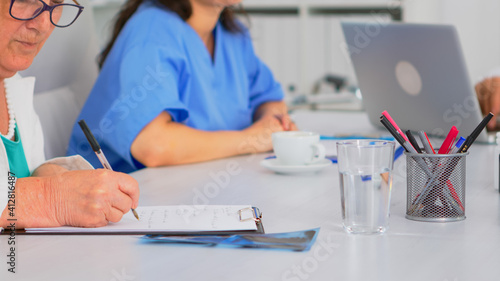 Close up of elderly woman doctor taking notes on clipboard while coworkers discussing in background  writing on laptop during brainstorming. Profesional teamworker having medical meeting