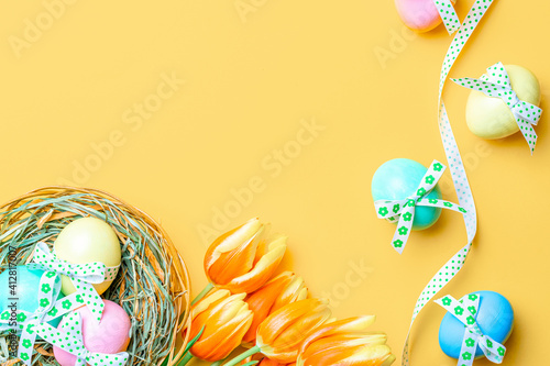 Easter frame. Colorful egg with tape ribbon, spring tulips, feathers on pastel yellow background in Happy Easter decoration. Festive design in flat lay.
