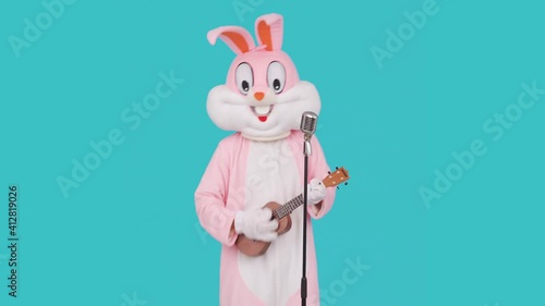 Singer guitarist is playing music by ukulele or Hawaiian guitar, sings song to retro vintage classic microphone. Easter bunny or rabbit or hare celebrates Happy easter.
