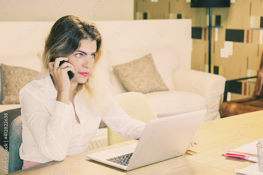 Serious designer discussing project with customer on phone. Young business woman talking on cell in coworking space, sitting at table with laptop and blueprints. Communication or multitasking concept Serious designer discussing project with customer on phone. Young business woman talking on cell in coworking space, sitting at table with laptop and blueprints. Communication or multitasking concept