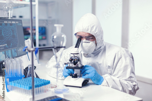 Analyzing virus in microbiology lab using microscope wearing ppe suit and glasses. Virolog in coverall during coronavirus outbreak conducting healthcare scientific analysis.