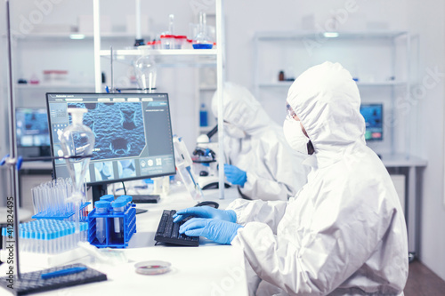Research scientist typing information obtained working in modern laboratory. Medical engineer using computer during global pandemic with coronavirus dressed in coverall.