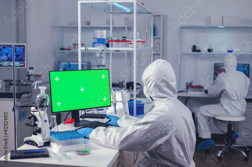 Doctor in protection suit against covid19 delevoping cure using computer with green screen dressed in ppe. Team of microbiologists doing vaccine research writing on device with chroma key  isolated