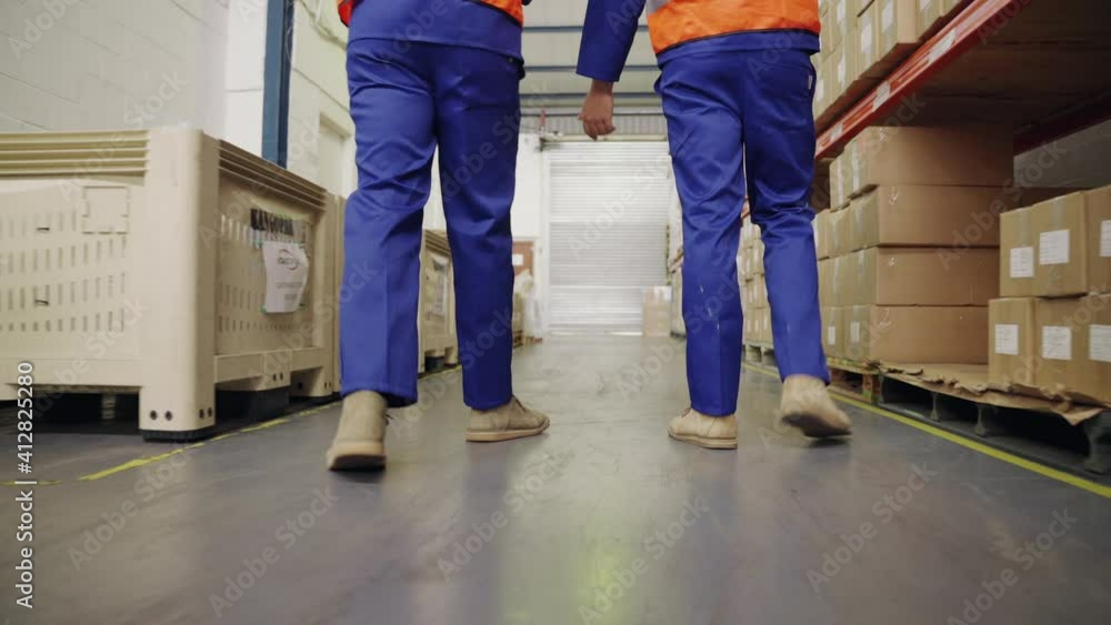 Low section of two male workers walking in the large distribution warehouse Low section of two male workers walking in the large distribution warehouse