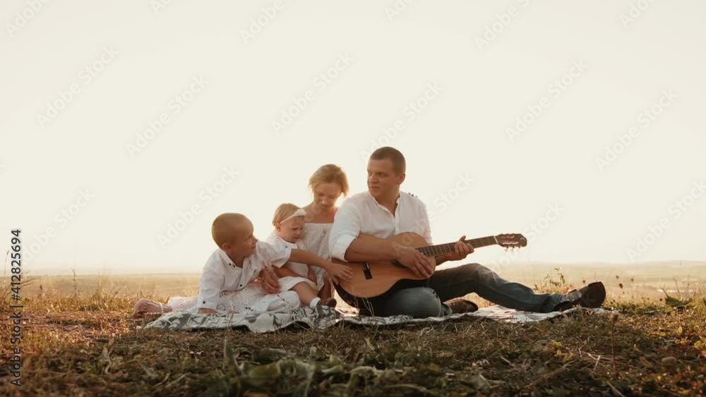 Friendly happy family of four sits at sunset and dad takes guitar to play and says something to son, who beats hand on it. Mom holds little obedient daughter on lap. Sun's rays break through people Friendly happy family of four sits at sunset and dad takes guitar to play and says something to son, who beats hand on it. Mom holds little obedient daughter on lap. Sun's rays break through people