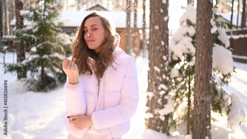 Winter sunny cold portrait in snowfall of beautiful young woman in a white warm outwear down jacket in backyard of country wooden house on the background of green spruce pine trees covered with snow Winter sunny cold portrait in snowfall of beautiful young woman in a white warm outwear down jacket in backyard of country wooden house on the background of green spruce pine trees covered with snow