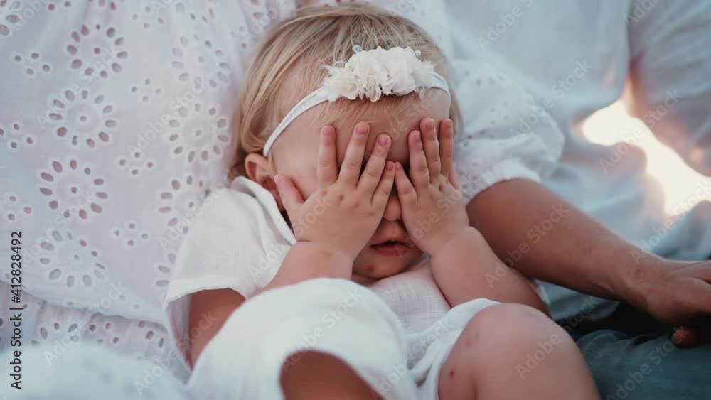 The little shy daughter sits leaning on her mother and closes her eyes with chubby tiny palms and then opens and smiles. Pretty cute girl in a white dress and a headband with flowers on her head The little shy daughter sits leaning on her mother and closes her eyes with chubby tiny palms and then opens and smiles. Pretty cute girl in a white dress and a headband with flowers on her head
