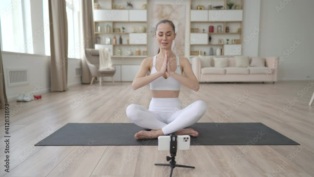 Young multi ethnic yoga coach, practicing breathing yoga pranayama on living room, recording online stream training tutorial for personal video blog, webinar web cam education entertainment concept Young multi ethnic yoga coach, practicing breathing yoga pranayama on living room, recording online stream training tutorial for personal video blog, webinar web cam education entertainment concept